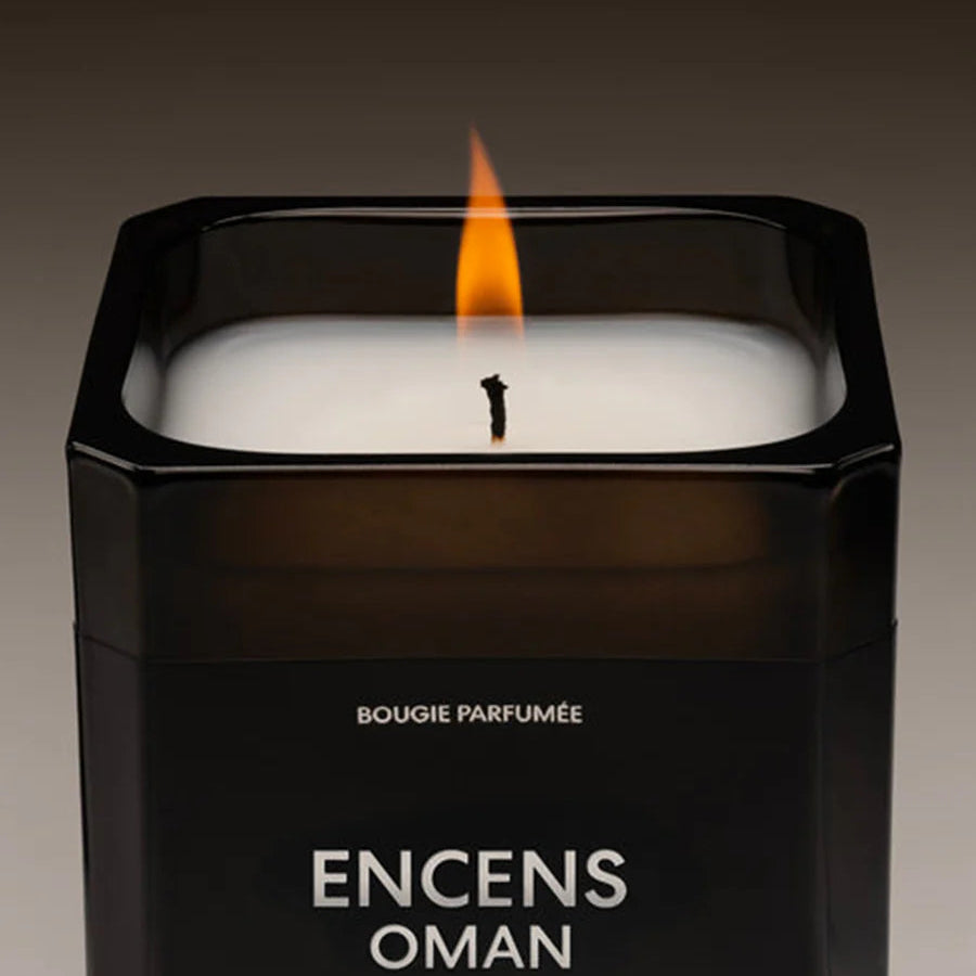Candle in a black container labeled 'Encens Oman' with a visible flame.